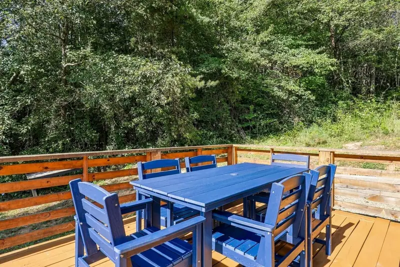 Blue outdoor dining table with six chairs on the cedar deck, with woods beyond the railing